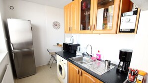 A compact kitchen with a stainless steel refrigerator, a washing machine, wooden cabinets, a black countertop, a sink, and a coffee maker. - Small one room apartment with balcony (Humenné)