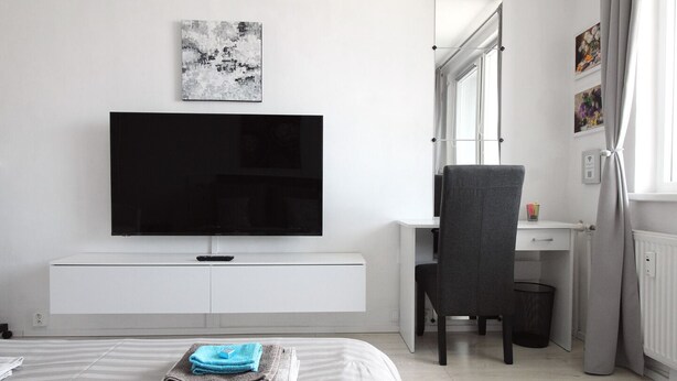 A modern bedroom with a large flat-screen TV mounted on a white wall, a grey chair, a desk with a computer, and a bed with neatly folded towels. - Small one room apartment with balcony (Humenné)