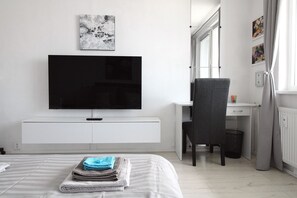 Une chambre moderne avec un grand téléviseur à écran plat fixé à un mur blanc, un fauteuil gris, un bureau avec un ordinateur et un lit avec des serviettes soigneusement pliées.