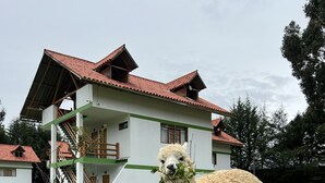 A white building with green trim and a red roof, surrounded by a grassy area with two alpacas grazing. - RESORT ALAPA (San Jerónimo de Tunán)