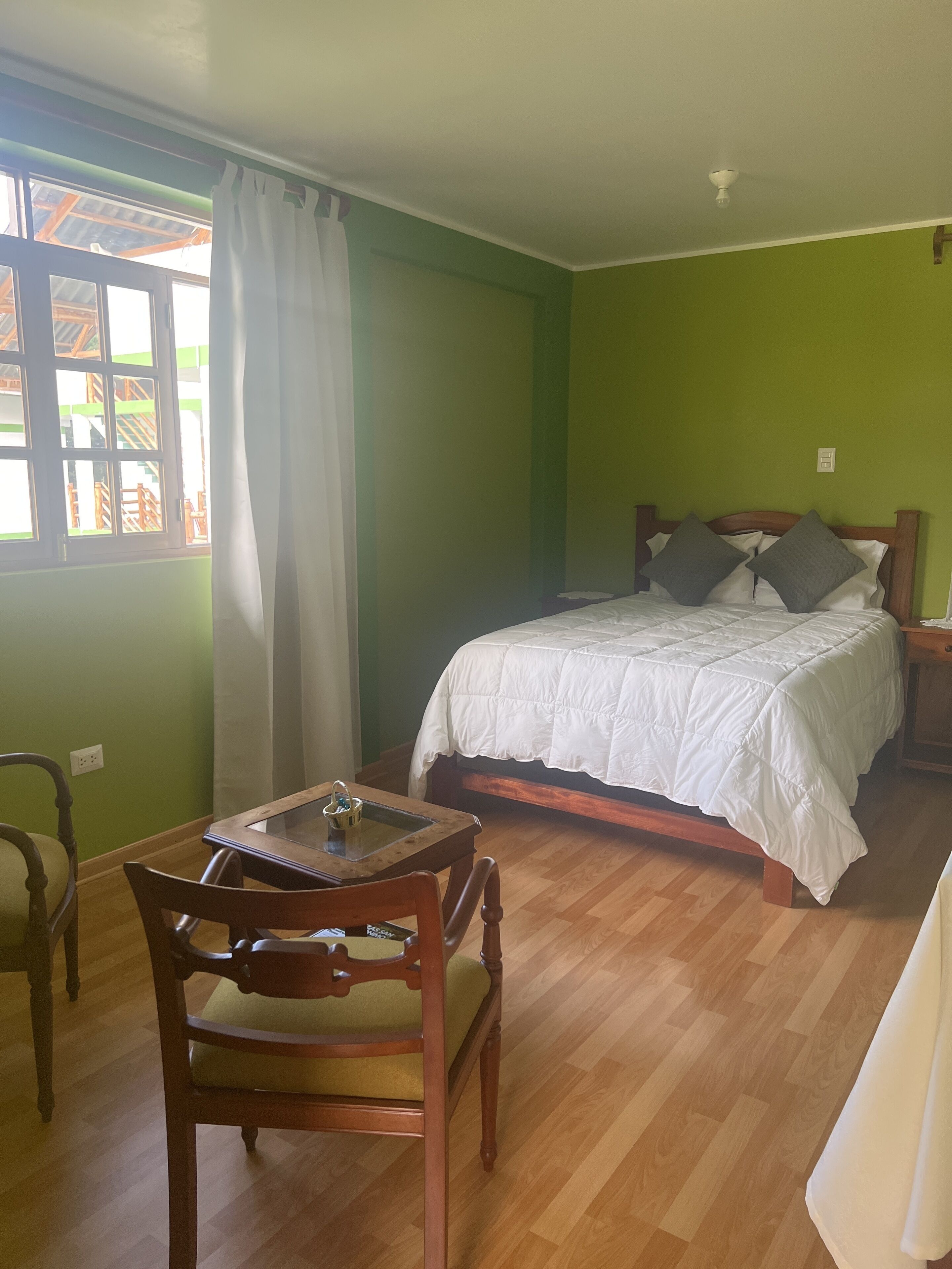 Una habitación con cama, una mesa con bandeja, una silla y un ventanal con cortinas.