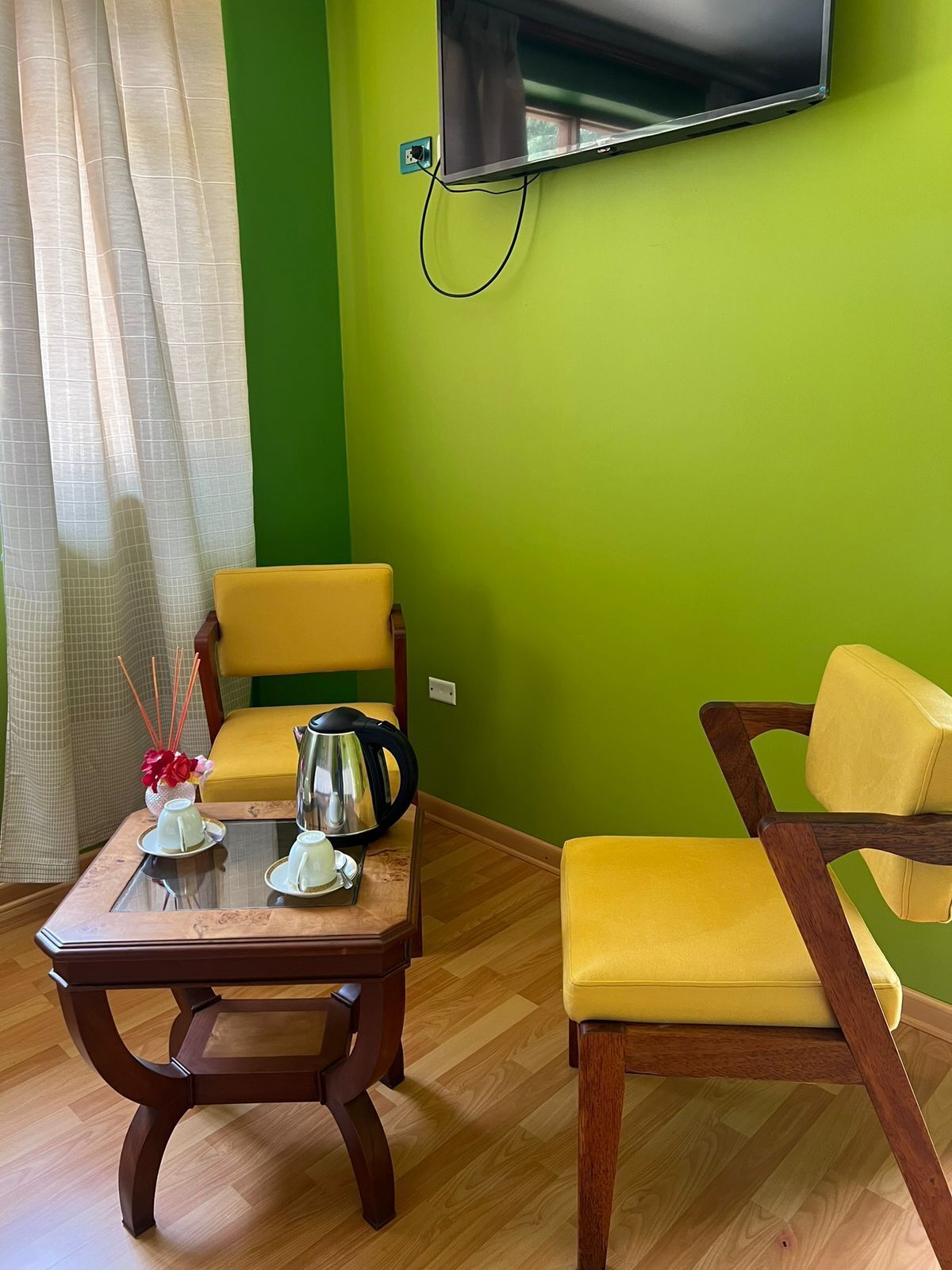 Una habitación con una pared verde, un televisor de pantalla plana montado en ella, una mesa de centro de madera con teteras y tazas, dos sillas amarillas y un suelo de madera.