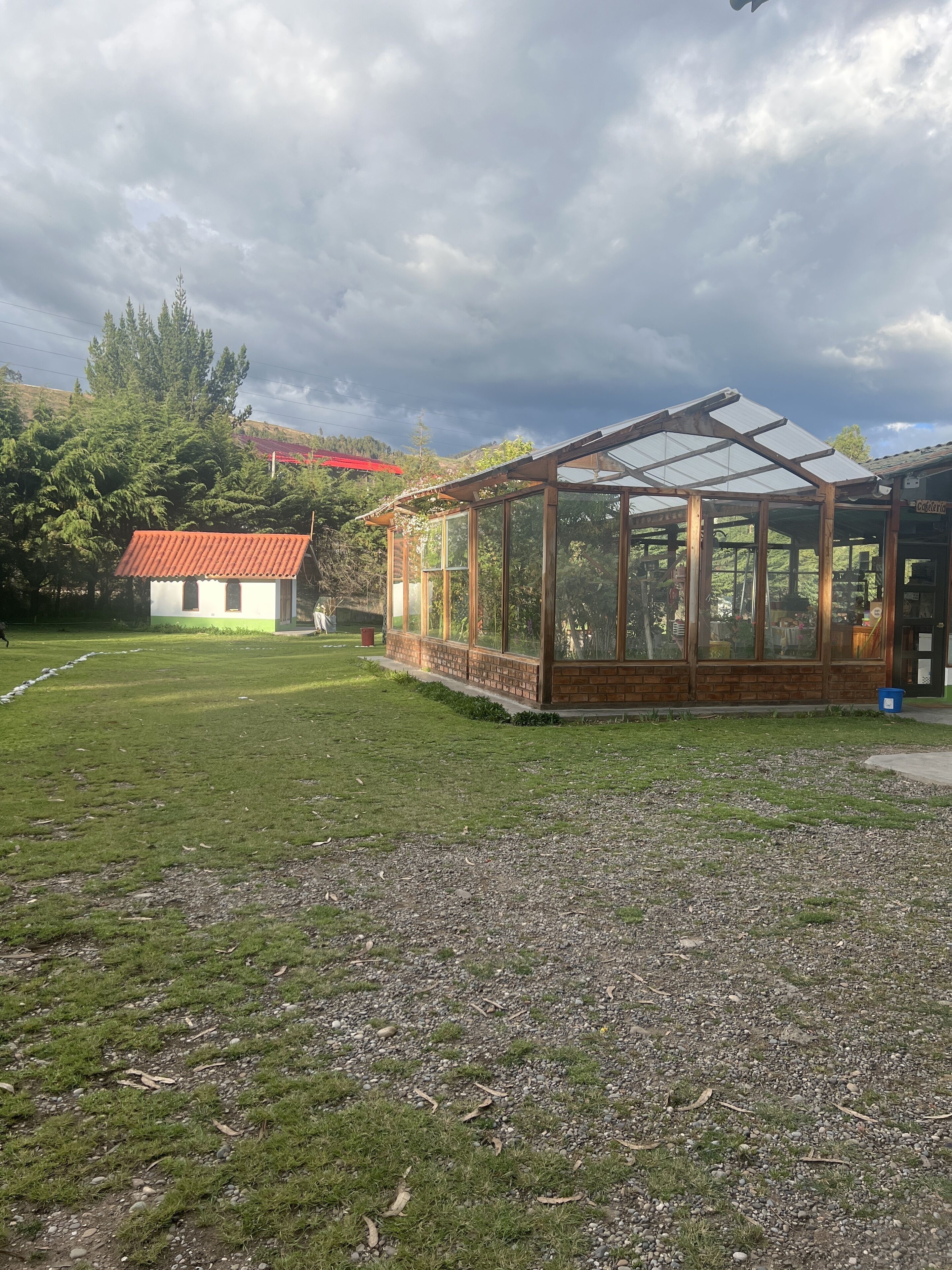 A conservatory with a glass roof, surrounded by a grassy area and a small building with a red roof.