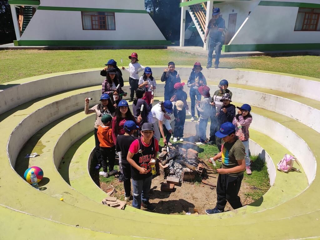 A group of people gathered around a campfire in a circular outdoor area with steps leading up to a building in the background.