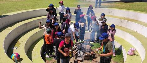 A group of people gathered around a campfire in a circular outdoor area with steps leading up to a building in the background.