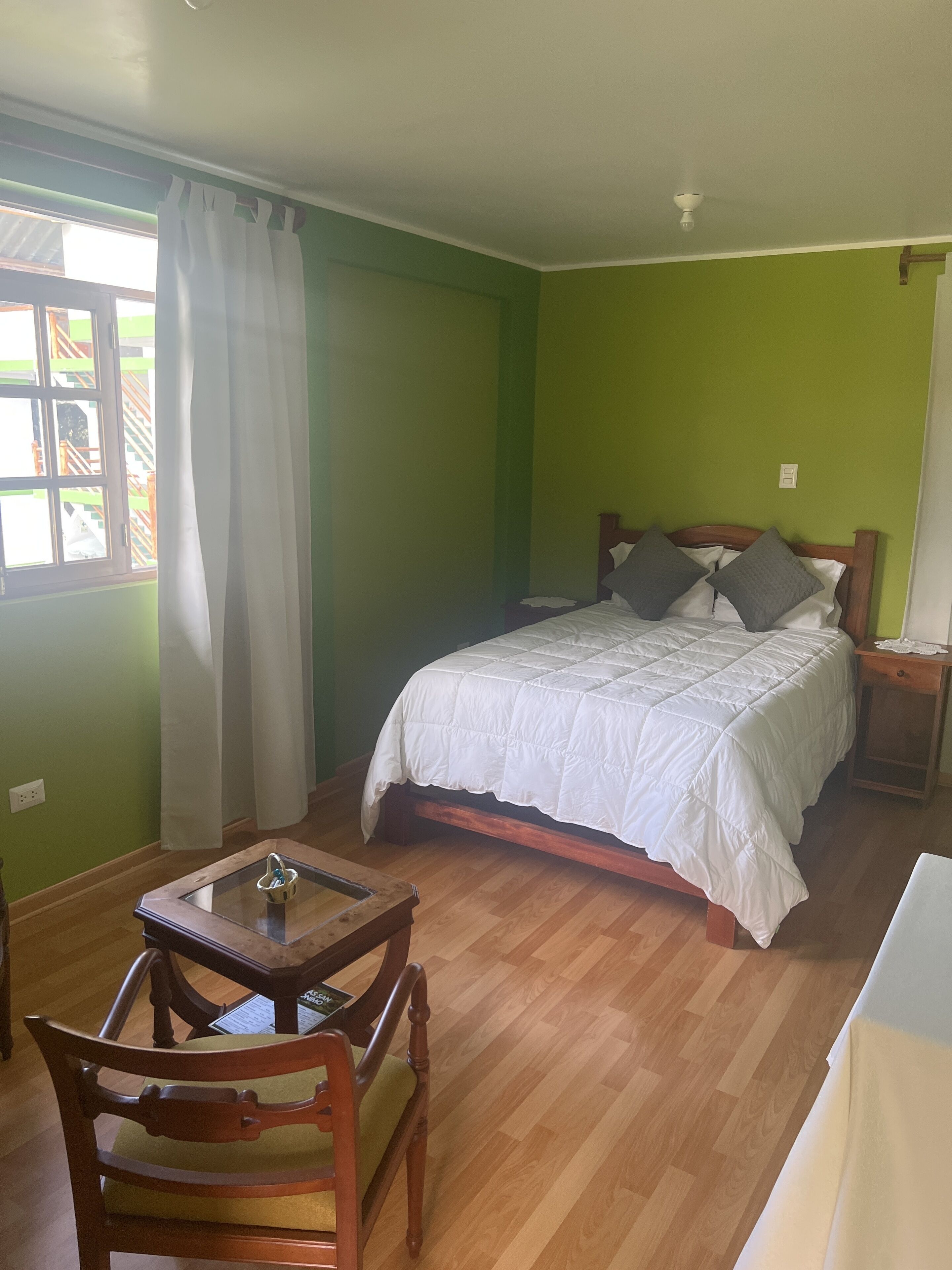 Un dormitorio con cama, mesita de noche, silla, mesita pequeña y ventana con cortinas.