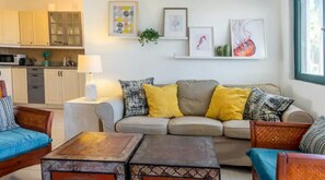 Un salon comprenant un canapé, deux fauteuils, une table basse et une table d’appoint avec une lampe. Au fond, on aperçoit une cuisine avec des placards et un four à micro-ondes.