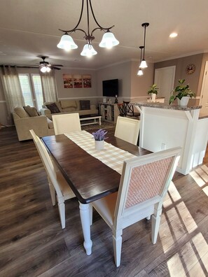 A dining area with a wooden table and white chairs, a kitchen in the background, and a ceiling fan. - Relax with your family or friends at this peaceful place to stay . (Navarre)