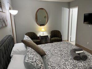 Une chambre d’hôtel avec un lit, deux fauteuils, une petite table avec une lampe, un miroir et un téléviseur fixé au mur.