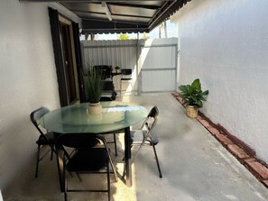 Une terrasse couverte avec une table en verre et quatre chaises, une plante en pot et un sol en béton.