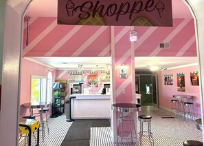 Interior de sorveteria com esquema de cores rosa e branco, balcão com cardápio e vários bancos.