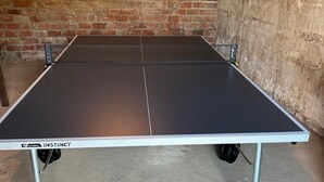 A ping pong table with a white border and the brand name 'Instinct' on the side, placed in a room with a brick wall and concrete floor.