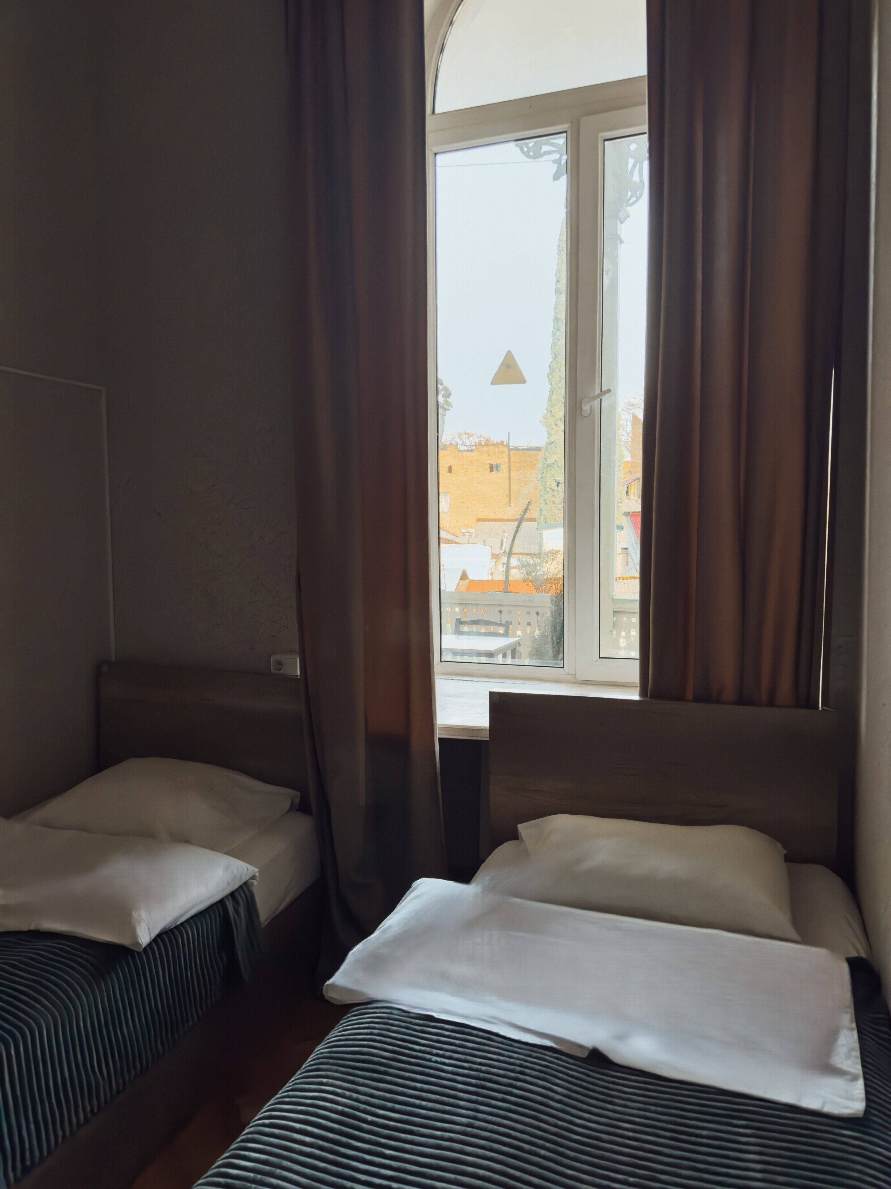 A hotel room with two single beds, a window with curtains, and a view of buildings outside.