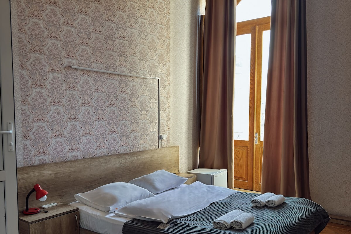 A hotel room with a wooden headboard, a bed with white linens, a nightstand with a lamp, and a view through a window with curtains.