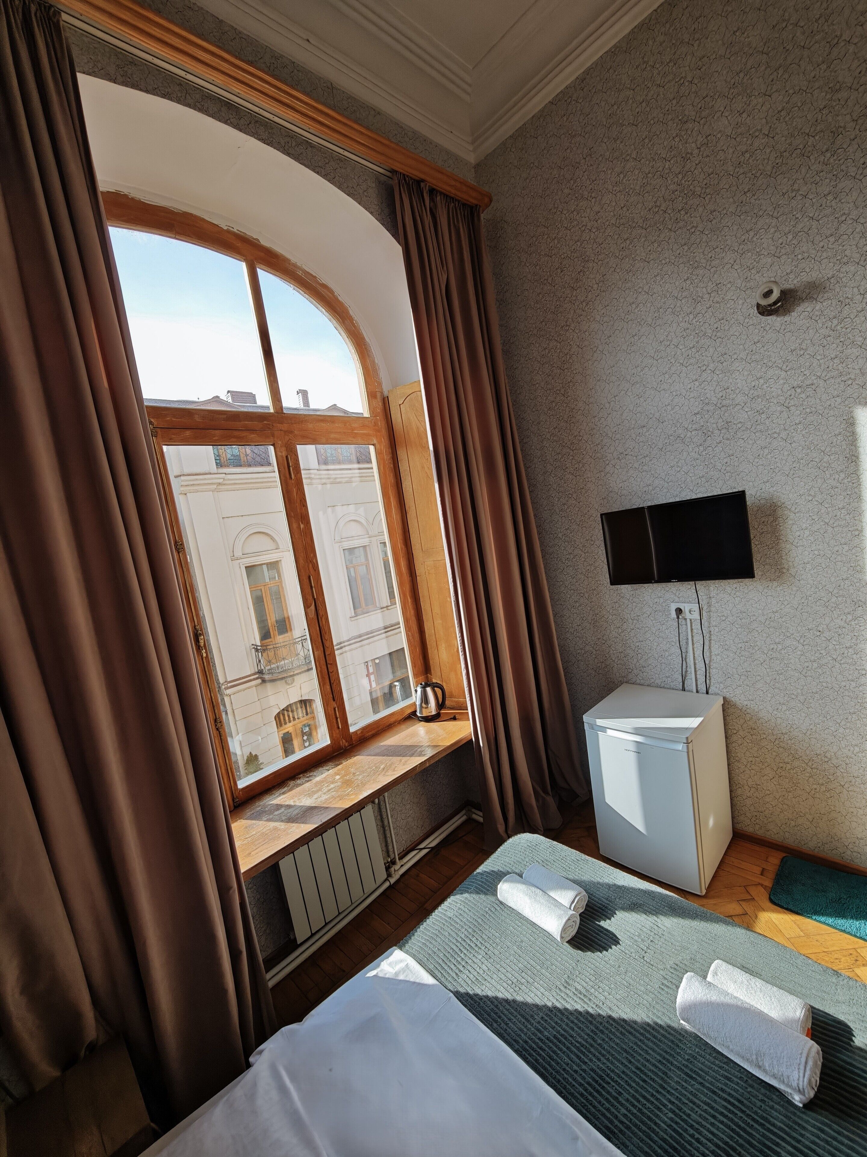 A hotel room with a large window, a bed with a green blanket, a small white heater, and a flat-screen TV mounted on the wall.