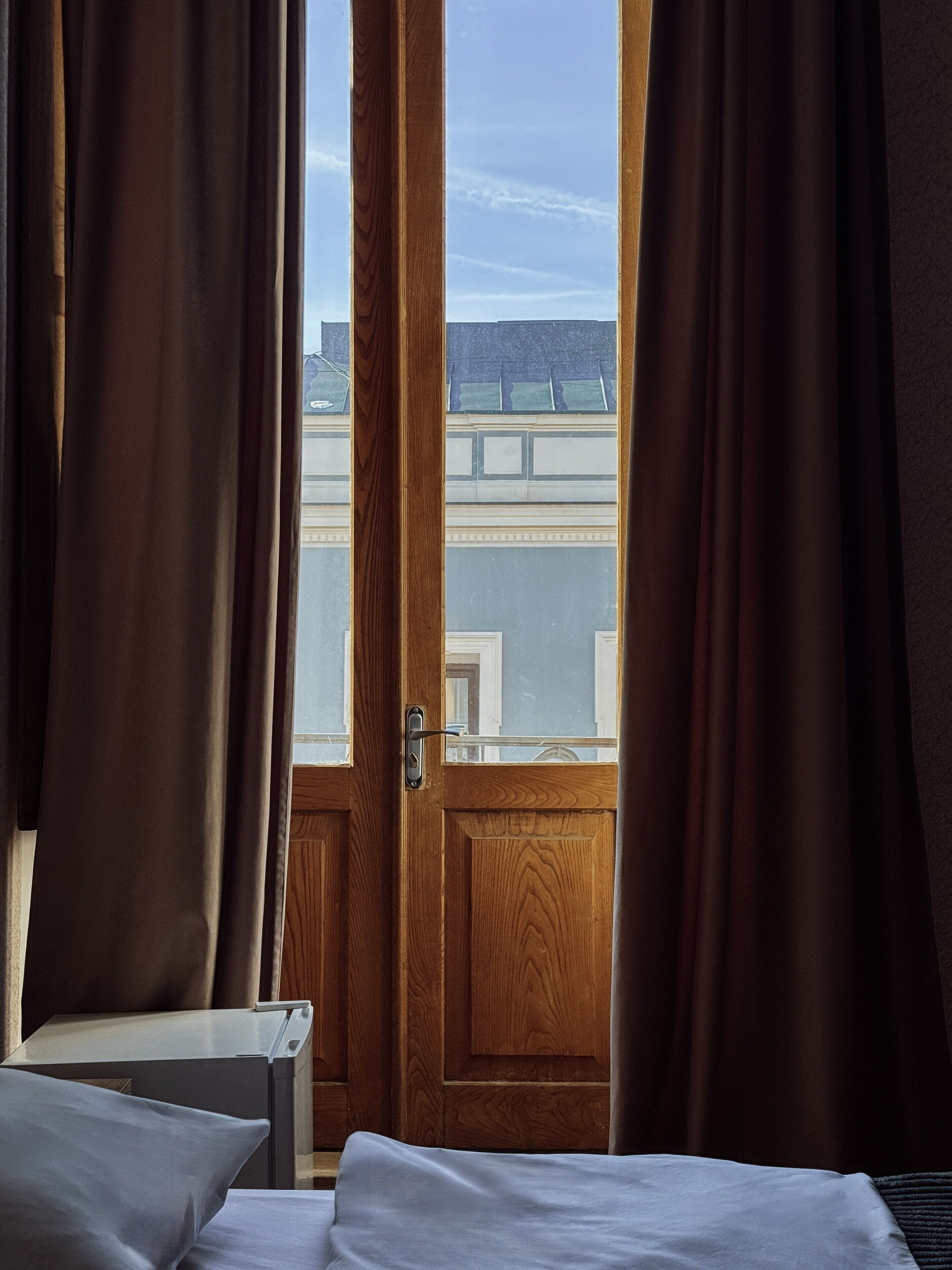 Una camera d'albergo con una porta in legno, vista sugli edifici dalla finestra e un letto con biancheria bianca.