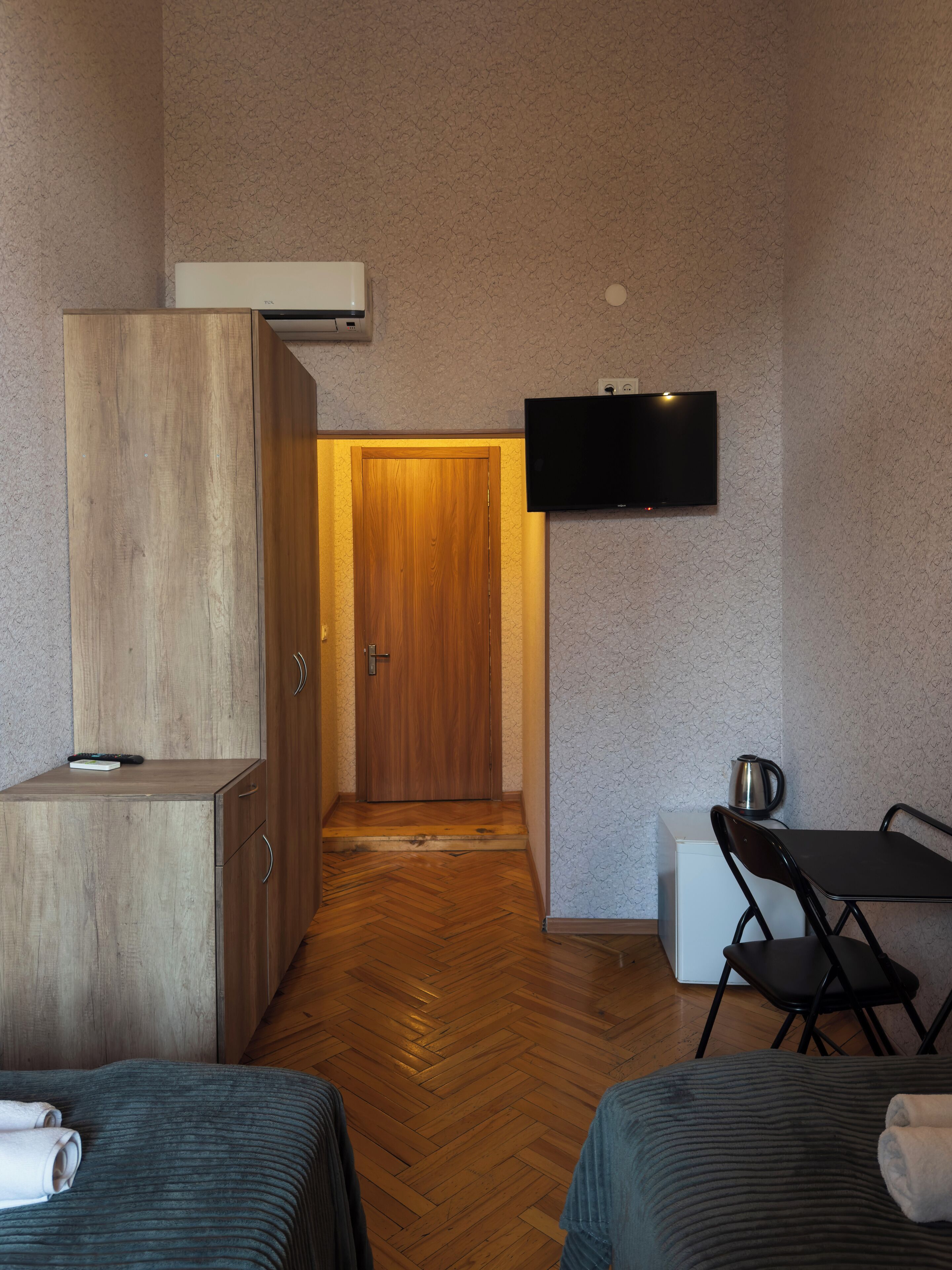 Une chambre d’hôtel avec une armoire en bois, un téléviseur fixé au mur, un petit coin cuisine, un plancher en bois et deux lits avec des serviettes.