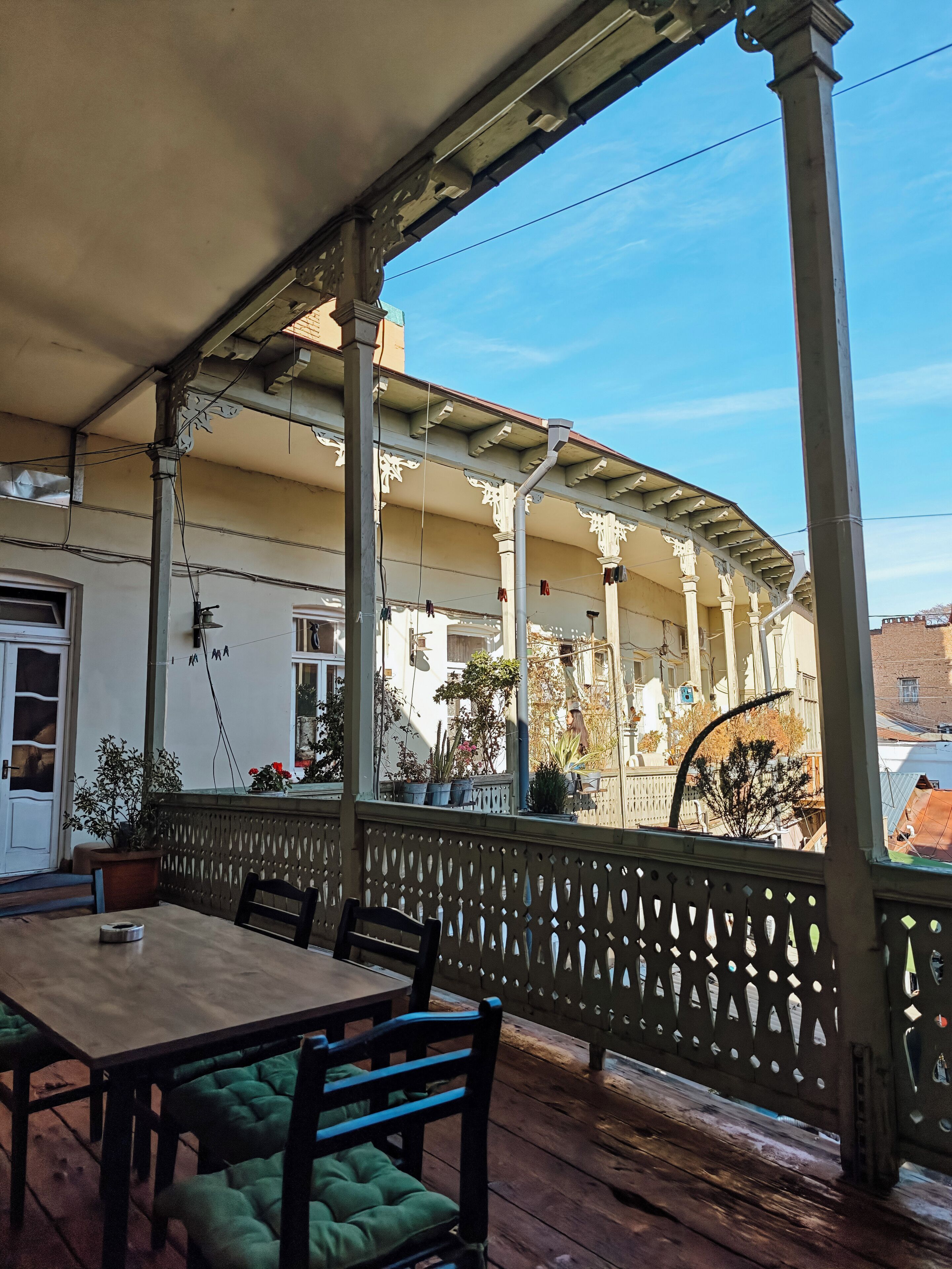 Un patio exterior con una mesa y sillas de madera, un área de estar cubierta con barandilla decorativa, y vistas a un edificio con columnas y plantas en macetas.