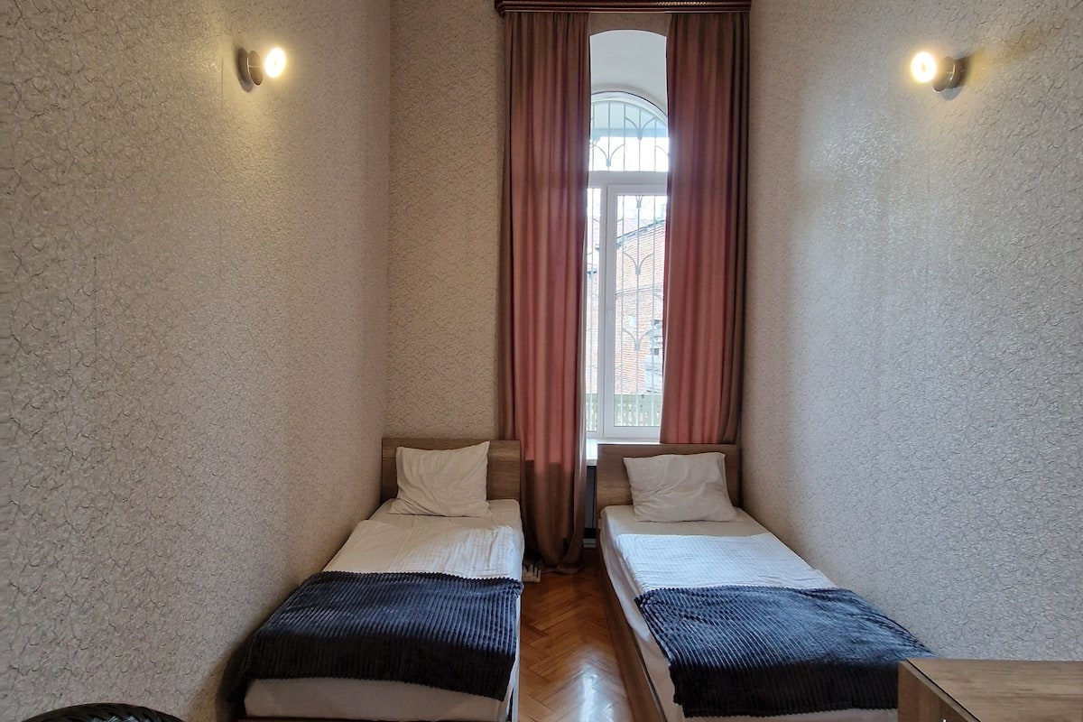 A small, well-lit room with two single beds, a wooden floor, a chair, and a cabinet.