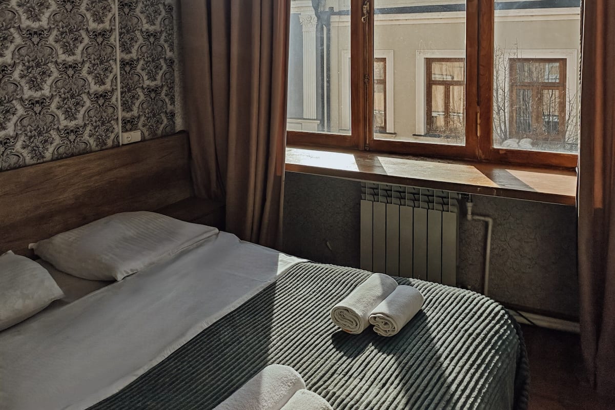 A hotel room with a large window, a bed with white linens, a radiator, and a patterned wallpaper.