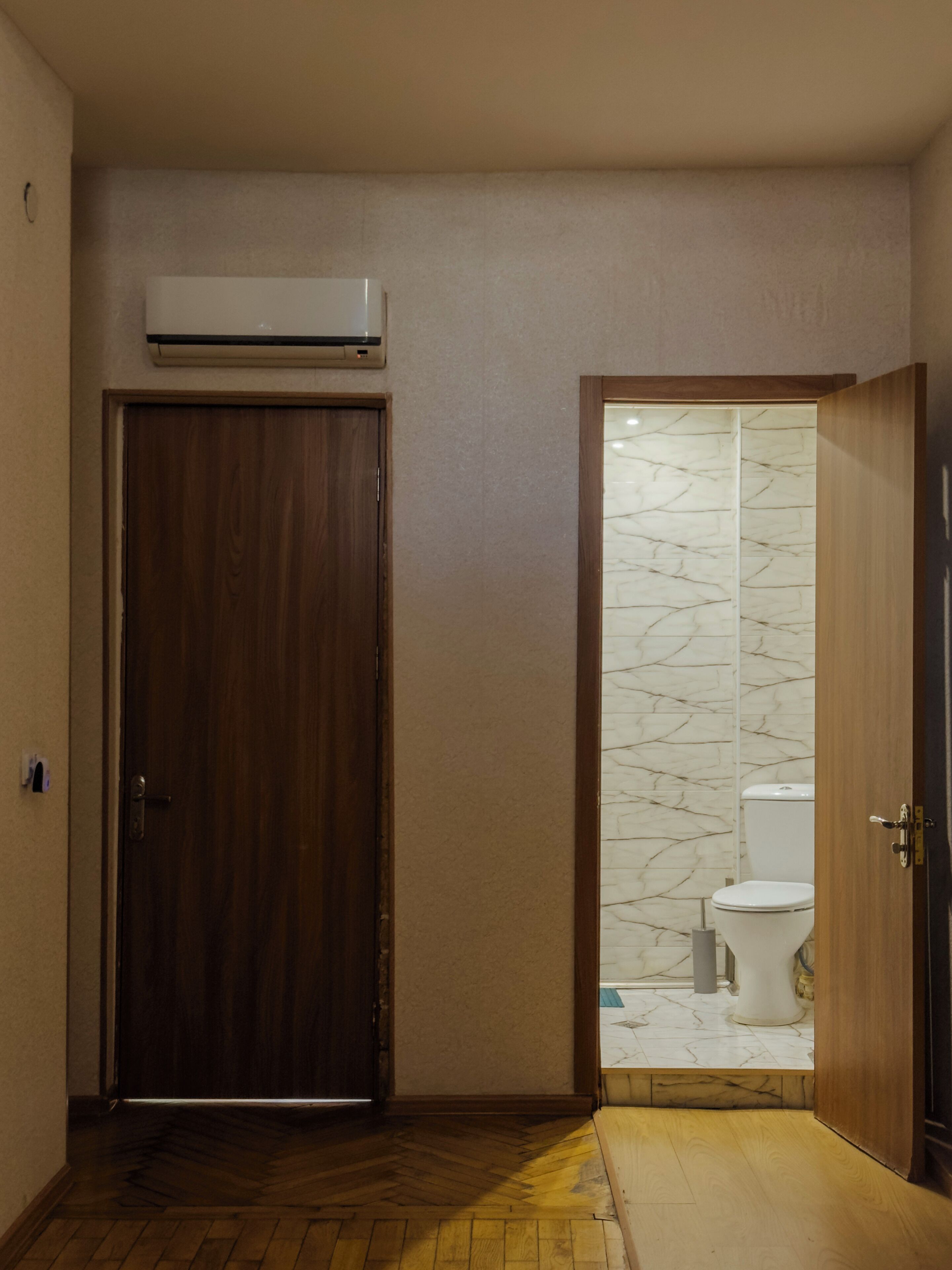 Un couloir avec une porte en bois, une salle de bain avec un mur en marbre et des toilettes visibles à travers une cloison en verre.