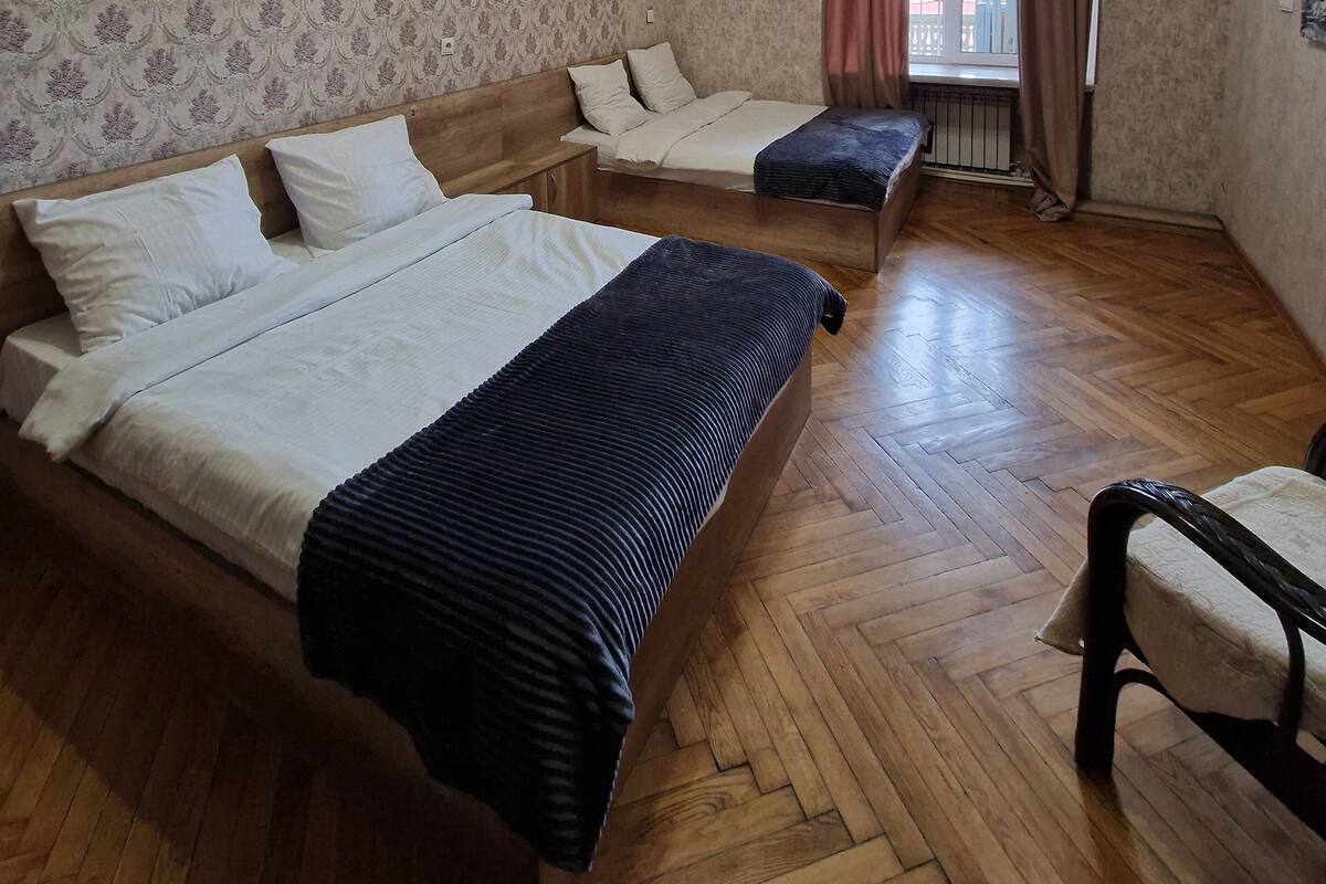 A room with two beds, a wooden floor, a patterned wallpaper, a window with curtains, and a chandelier.