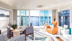 A modern living room with a large window overlooking a body of water, a dining area with a table and chairs, and a seating area with a sofa and armchairs. - New Floating Luxury: Unique Houseboat Retreat (Orange Park)