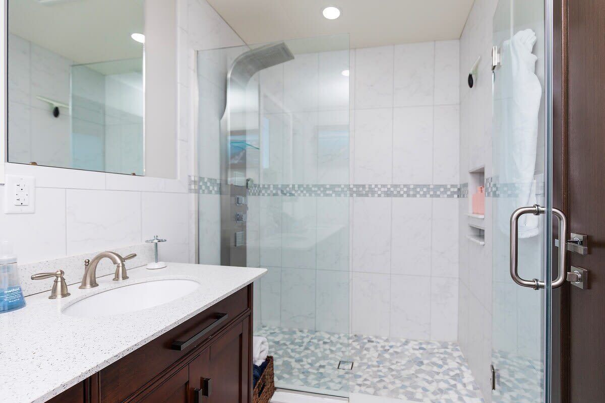 Un baño con ducha de vidrio, lavabo blanco sobre encimera de mármol y tocador de madera.