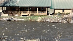 A lodge with a green roof, wooden deck, and a view of a river. - Rio Cabin - Serenity on the South Fork (South Fork)