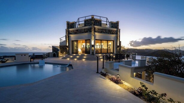 A modern two-story house with a swimming pool, outdoor seating, and a view of the ocean.