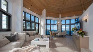 A spacious living area with a large sofa, a coffee table, and a dining table with chairs. There are large windows providing views of the outdoors.