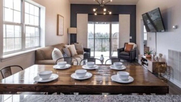 A dining area with a wooden table set for four, a sofa, and a TV mounted on the wall. - Gated, Luxe Mid-Century Modern Condo| Walk to golf & pools! (Branson)