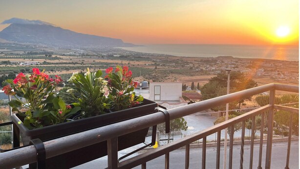 Ein Balkon mit Blick auf einen Sonnenuntergang über einer Küstenlandschaft, einschließlich eines Berges und einer Stadt in der Ferne.