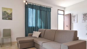 Ein Wohnzimmer mit einem beigefarbenen Ecksofa, einem einzelnen Sessel, blaugrünen Vorhängen und Kunstwerken an den Wänden.