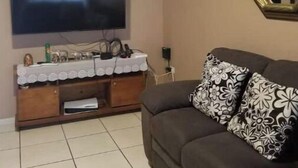 Ett vardagsrum med en platt-TV som är monterad på väggen, ett träinrett mediaskåp under TV:n och en mörkfärgad soffa med mönstrade kuddar.