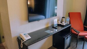 A hotel room with a flat-screen TV mounted on the wall, a desk with a telephone, a coffee maker, and a chair with an orange cushion. - Golden Sands Destination Resorts (Cebu Island)