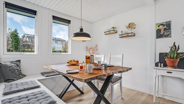 Ein stilvoller Essbereich mit einem Holztisch, weißen Stühlen und einer schwarzen Pendelleuchte. Zwei Fenster mit Jalousien, eine weiße Wand mit einer kleinen Ablage und ein Blumentopf auf einem weißen Schrank.