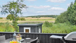 Eine Holzterrasse mit Tisch, der für eine Mahlzeit gedeckt ist, einem Grill und Blick auf ein Feld und Bäume.
