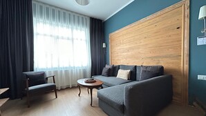 A modern living room with a grey sofa, a wooden coffee table, and a chair. There is a large window with sheer curtains, a wooden accent wall, and a lamp on the right. - Istanbul Gem: 3-Bedroom Apartment with Panoramic Terrace and Iconic Sea Views (İstanbul)