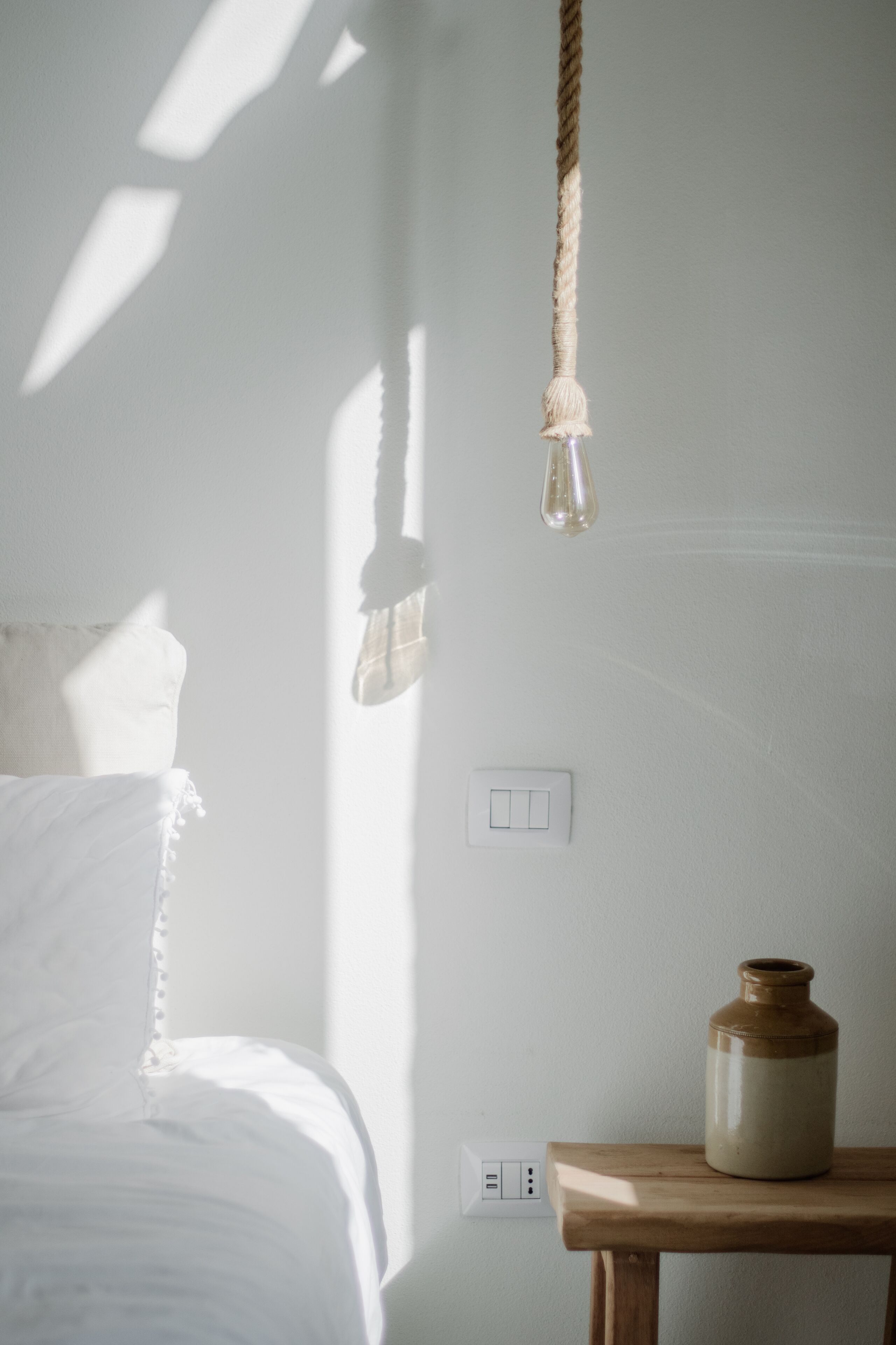 Una stanza con un letto, un comodino in legno, un lampadario a sospensione e una presa elettrica a muro.