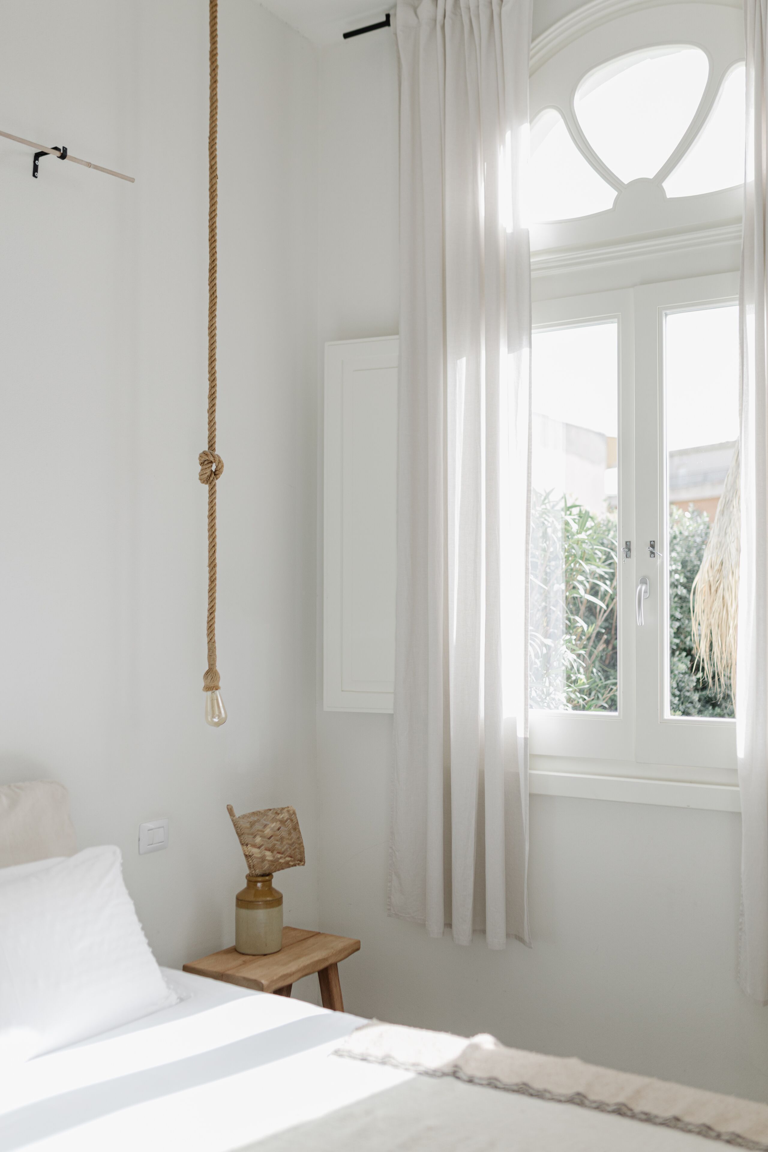 Une chambre à coucher avec un lit, une guirlande lumineuse suspendue, une petite table en bois avec une lampe et une fenêtre avec des rideaux.