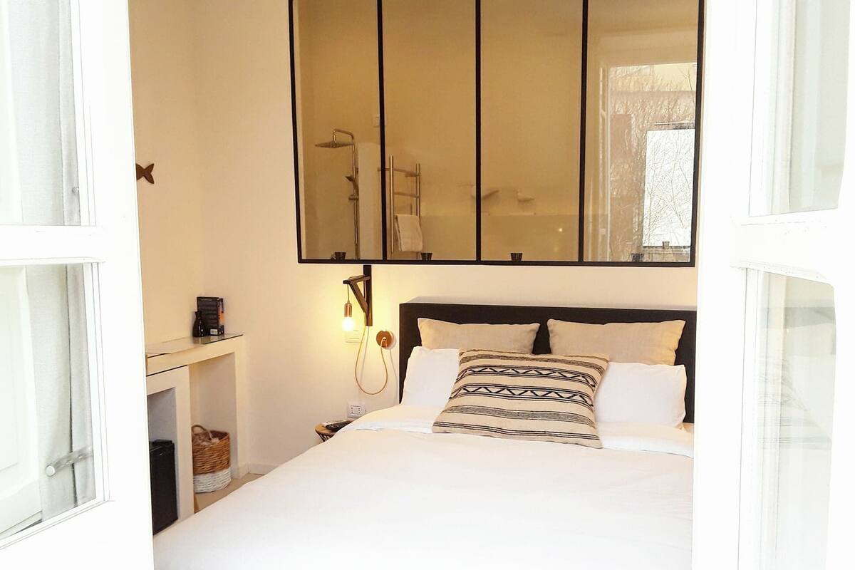 Een slaapkamer met een bed, nachtkastje met lamp, spiegel boven het bed en een raam met gordijnen.