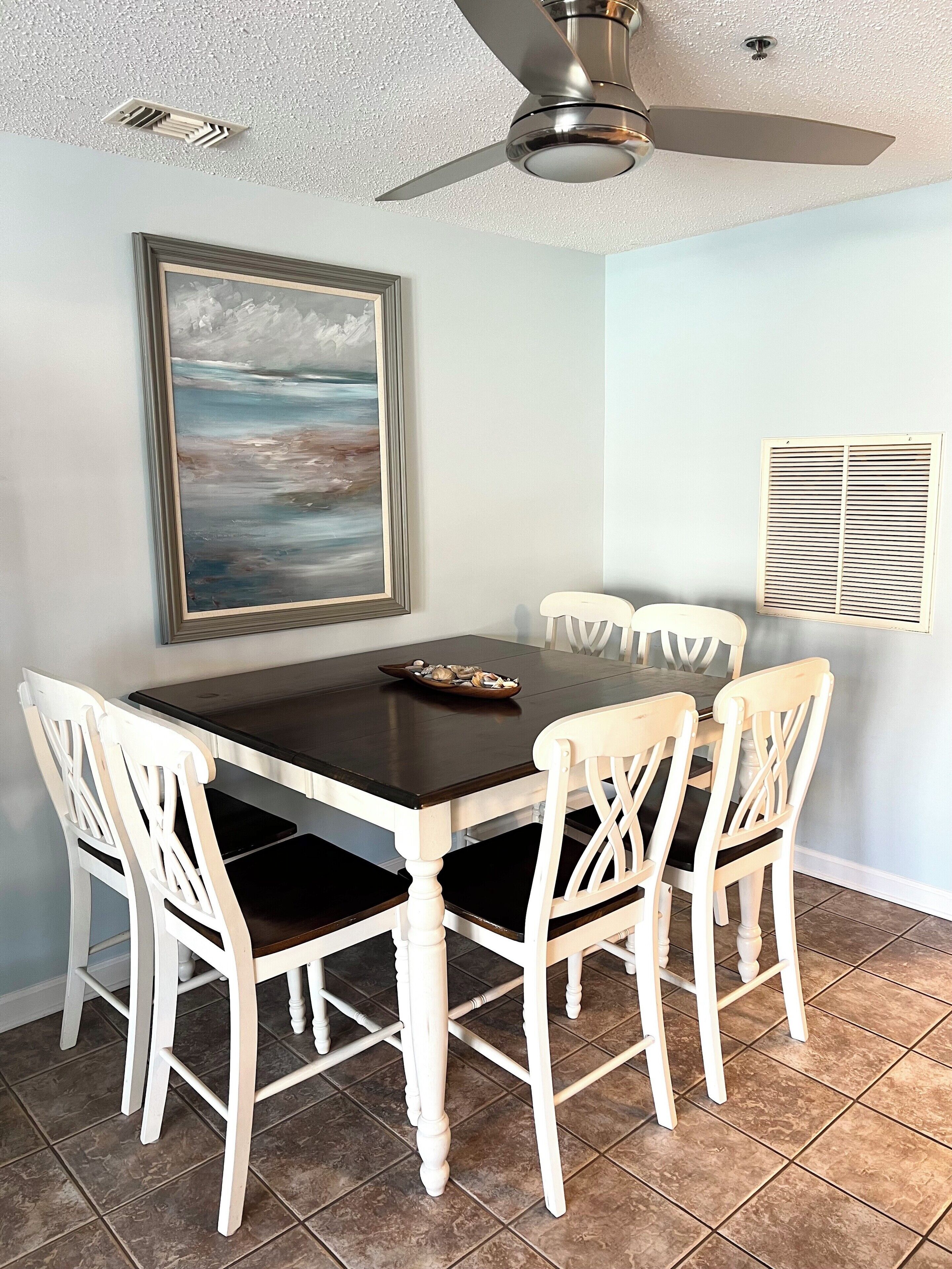 Un coin repas avec une table en bois foncé, des chaises blanches, un ventilateur de plafond, un tableau encadré et une fenêtre avec des stores.
