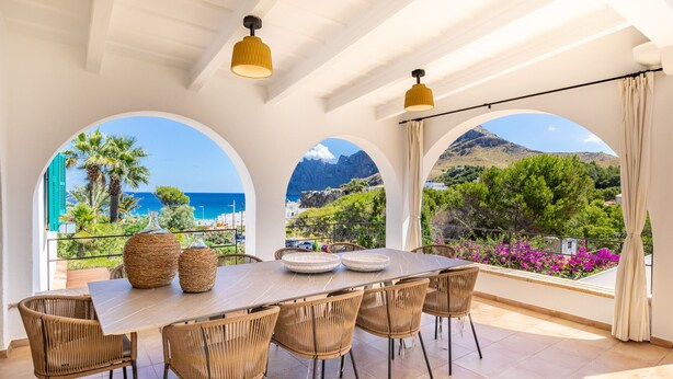 A dining area with a table and chairs, large windows with arched frames, and a view of the sea and mountains. - Cavall Bernat 14 - Majorca Villa by the beach (Cala Sant Vicenç)