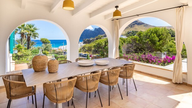A dining area with a large table, six wicker chairs, and two woven baskets. It features large arched windows with a view of the sea and mountains. - Cavall Bernat - Villa by the sea Majorca (Cala Sant Vicenç)