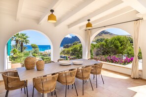 Un coin repas comprenant une grande table, six chaises en osier et deux paniers tressés. Il est agrémenté de grandes fenêtres en arc de cercle offrant une vue sur la mer et les montagnes.
