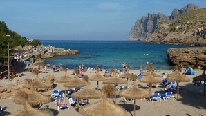 A beach with thatched umbrellas, sun loungers, and rocky cliffs in the background. - Cavall Bernat - Villa by the sea Majorca (Cala Sant Vicenç)
