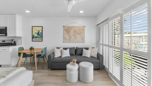 Una sala de estar moderna con un sofá gris, una mesa redonda de centro, y una zona de comedor con una pequeña mesa y sillas. Hay un ventilador de techo, un mapa del mundo en la pared y una ventana con persianas.