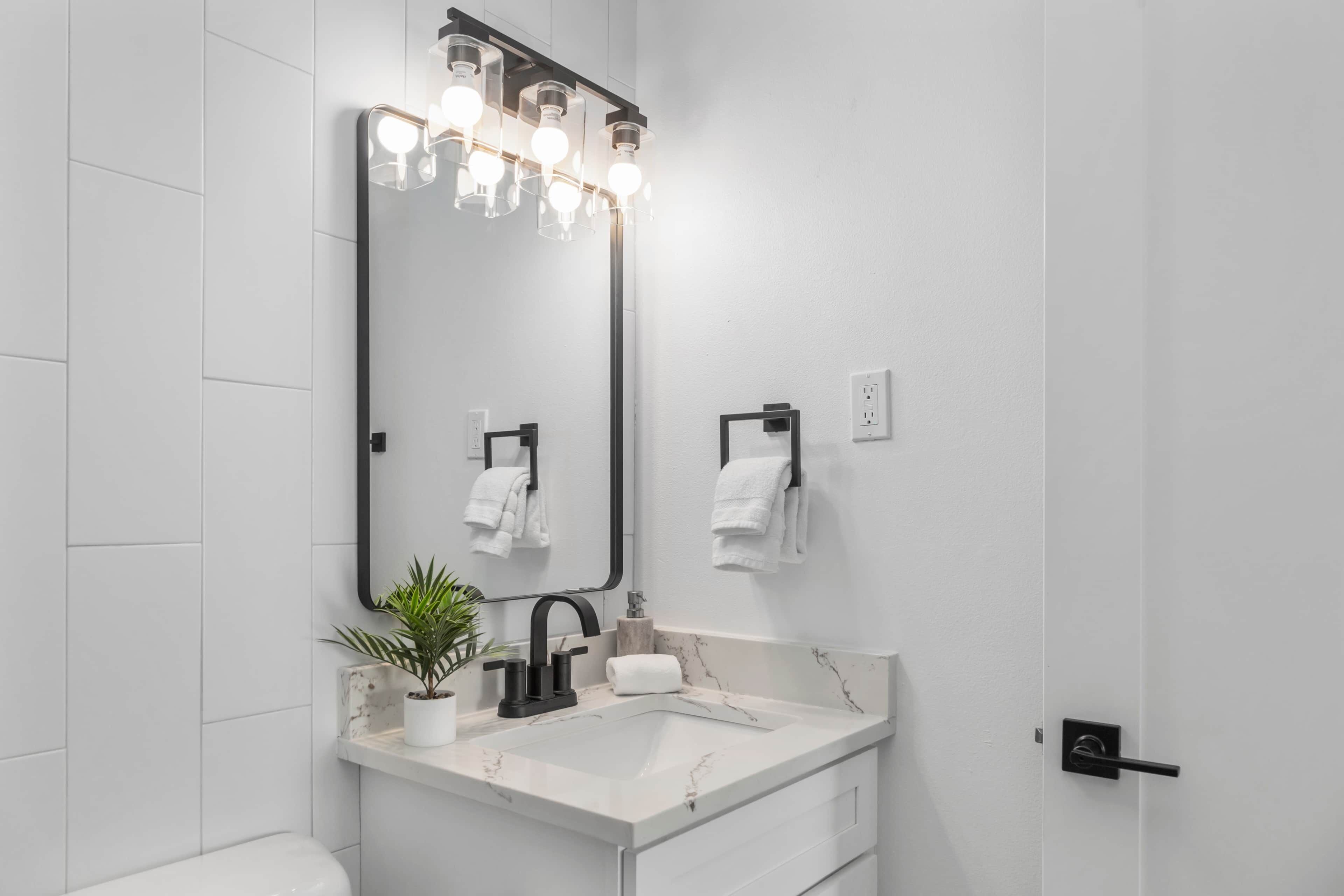 Casa de banho moderna com lavatório branco, torneira preta e espelho grande com iluminação. Toalhas penduradas em ganchos e uma planta em vaso no balcão.