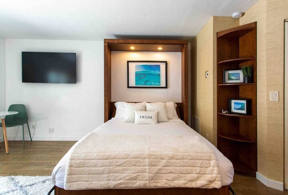 A neatly arranged bedroom with a bed, a framed picture above it, a small desk with a chair, and a wooden shelf with pictures and plants.